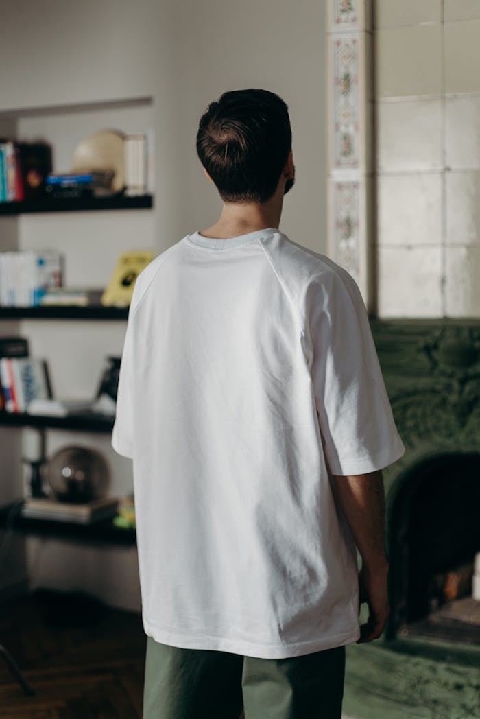 Adult male wearing a white t-shirt stands indoors facing a bookshelf.