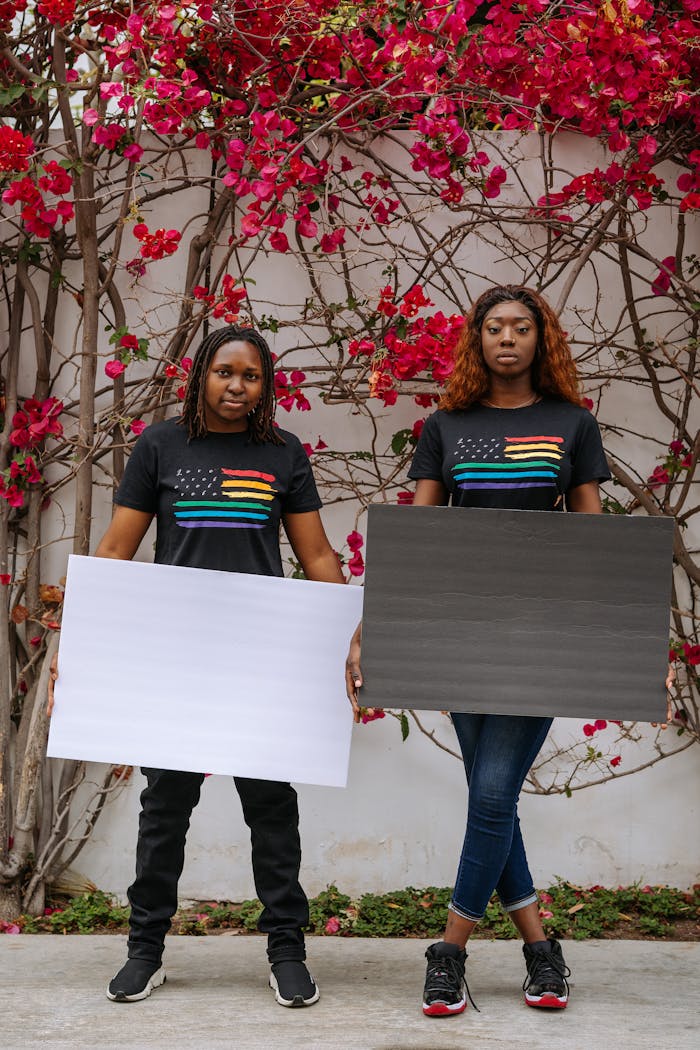 African American couple in rainbow flag shirts holding blank placards in front of vibrant flower wall.